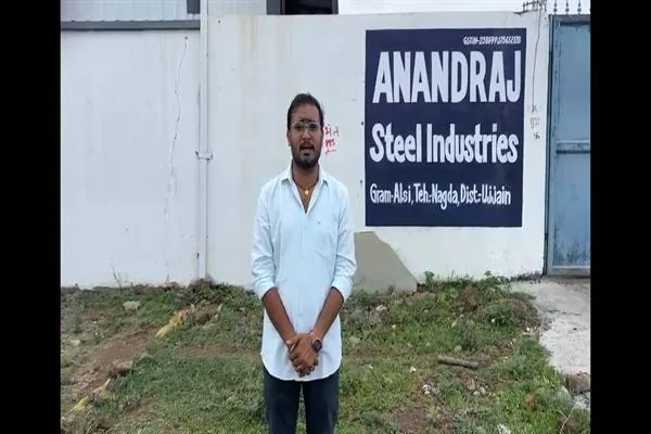सफेद कमीज पहने एक युवक आनंदराज स्टील इंडस्ट्रीज के बोर्ड के सामने खड़ा है