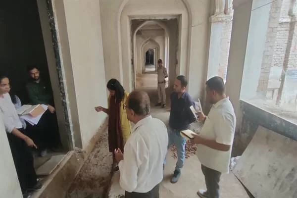 पुरानी इमारत के मेहराबदार गलियारे में खड़े अधिकारी और तकनीकी टीम के सदस्य निरीक्षण करते हुए, फर्श उखड़ा हुआ और दीवारों पर मरम्मत का काम दिख रहा है