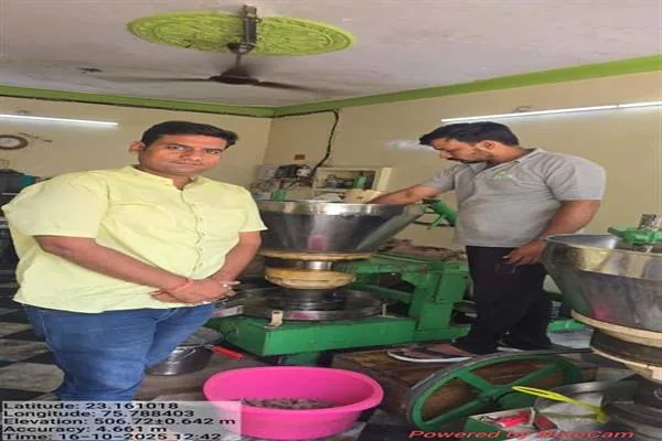 एक कमरे में लगी हरे रंग की कच्ची घानी तेल मशीन के पास खड़े दो पुरुष, मशीन के नीचे रखा गुलाबी टब और पास में धातु का बड़ा बर्तन।
